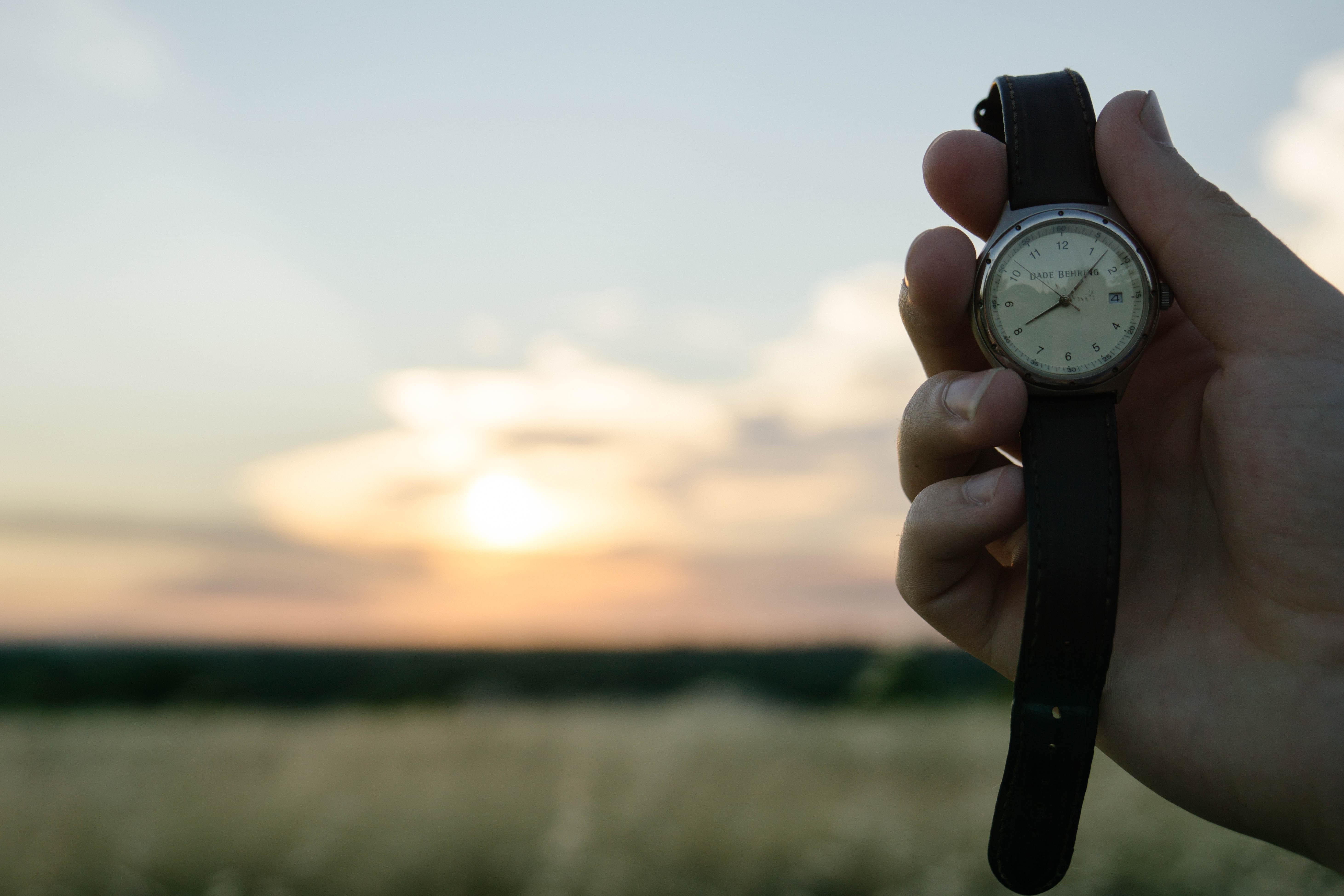 Mano sujetando un reloj de pulsera con correa de cuero con un paisaje de fondo. Foto de Wil Stewart en Unsplash